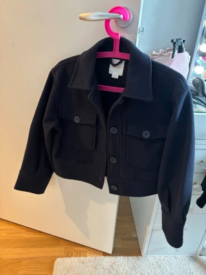 Jacka  - Stilren mörkblå jacka med stora knappar framtill. Två bröstfickor med lock och knappdetaljer ger en cool look. Jackan  har en boxig passform och är tillverkad i ett tjockare material som känns mjukt och värmande.