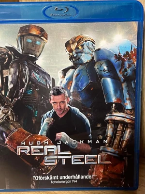 Blu-ray Real Steel - Blu-ray-film Real Steel med Hugh Jackman i huvudrollen. Omslaget visar robotar i futuristisk boxningsmiljö och filmen är på svenska, engelska och flera andra språk. Perfekt för dig som gillar action och sci-fi med robotar och spänning.