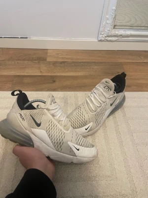 Nike Air Max 270 vita sneakers - Nike Air Max 270 sneakers i vitt med grå och svarta detaljer. Skorna har en luftig mesh-överdel, synlig Air-enhet i hälen och klassisk Nike-logga på sidan och tån. Snygg och sportig design som passar till streetwear och casual outfits.