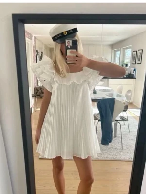 Vit plisserad kortklänning med volang - Supersöt vit kortklänning med plisserad kjol och stora volangdetaljer. Perfekt till student eller skolavslutning❤️