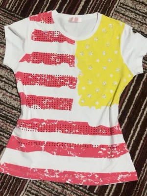 Färgglad t-shirt med stjärnor och ränder - Unik t-shirt med vita, röda och gula partier. Framsidan har röda ränder med slitna detaljer och paljetter samt ett gult fält med vita stjärnor. Kortärmad och figurnära passform, perfekt för dig som gillar färg och mönster.