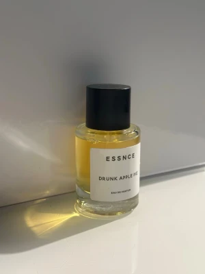 ESSNCE Drunk Apple Pie parfym - ESSNCE Drunk Apple Pie är en Eau de Parfum i en rund glasflaska med svart plastlock. Doften har en varm gul färg och etiketten är vit med minimalistisk text. Perfekt för dig som gillar fruktiga och söta dofter.