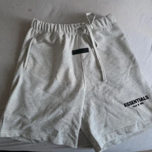 Gråa Essentials shorts med snörning - Säljer ett par ljusgrå shorts från Essentials Fear of God med snörning i midjan och tryckt logga på benet. Shortsen har elastisk midja och sidofickor, perfekta för en avslappnad stil.