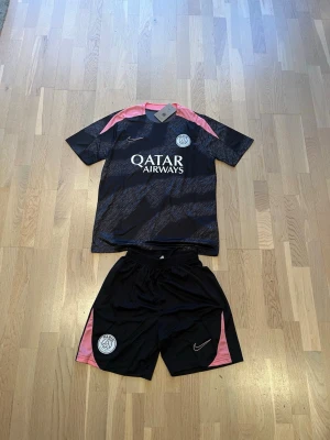 PSG träningsset svart/rosa Nike - Säljer ett PSG träningsset från Nike med svart och rosa design. Tröjan har Qatar Airways-tryck och PSG-logga på bröstet, samt rosa detaljer på axlarna. Matchande shorts med PSG-logga och rosa paneler på sidorna. Perfekt för fotbollsträning eller gymmet. Storlek S