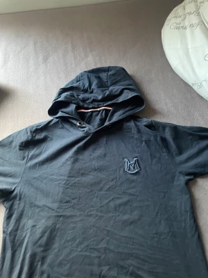 Svart T-shirt  från Moncler - Snygg svart t shirt med luva,  från Moncler med stor logga tryckt längs ryggen och en mindre broderad logga på bröstet. Tillverkad i mjukt bomullsmaterial och har en klassisk huva med dragsko. Perfekt för en avslappnad och trendig look.