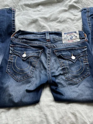 Blå True Religion jeans med kontrastsömmar - Säljer ett par blå jeans från True Religion med ikoniska kontrastsömmar och stora ficklock bak. Jeansen har silverfärgade knappar och detaljer samt en patch med logga bak i midjan. Klassisk femficksmodell i slitstark denim.