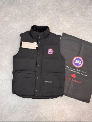 Svart dunväst från Canada Goose - Snygg svart dunväst från Canada Goose med klassisk logga på bröstet. Västen har hög krage, två stora framfickor och stängs med tryckknappar. Perfekt för lager-på-lager och håller dig varm med sin vadderade design.