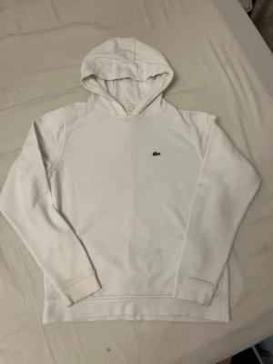 Vit hoodie från Lacoste - Snygg vit hoodie från Lacoste med klassisk krokodillogga broderad på bröstet. Hoodien har en enkel och stilren design med långa ärmar och mjukt material, perfekt för en avslappnad look.