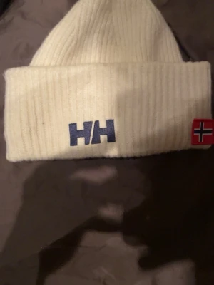 Vit ribbad mössa från Helly Hansen - Säljer en stilren vit ribbstickad mössa från Helly Hansen med uppvikt kant. Mössan har HH-logga framtill och en norsk flagga på sidan. Perfekt accessoar för kalla dagar och enkel att matcha med olika outfits.