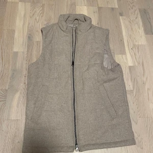 Beige dunväst med hög krage - Stilren beige dunväst från John Henric med hög krage och dragkedja framtill. Västen har två snedställda fickor och är tillverkad i ett mjukt tyg med diskret struktur. Perfekt för lager-på-lager och enkel att matcha med olika outfits. Säljer denna då den har blivit för liten. Västen är aldrig använd.