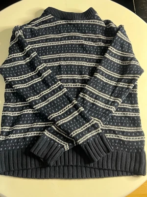 Mörkblå stickad tröja med mönster - Mysig mörkblå stickad tröja med vita och ljusblå ränder och småmönster. Tröjan har ribbstickade muddar vid ärmslut och nederkant samt en klassisk rund halsringning. Perfekt för kalla dagar och enkel att matcha med jeans.