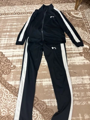 Svart träningsset med vita ränder - Snyggt svart träningsset från Puma med vita ränder längs ärmar och ben. Setet består av en zip-jacka och matchande byxor, båda med Puma-logga. Perfekt för träning eller chill. Materialet är troligtvis polyester för en sportig känsla.