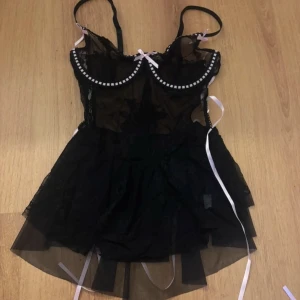 Svart babydoll - Svart babydoll med spetsdetaljer och rosa band. Babydollen har justerbara axelband och en matchande trosa & strumpeband medföljer. Designen inkluderar snörning i sidorna och små rosetter. Endast testat allt en gång, trosorna med andra trosor under. Strl M på allt, babydollen & trosan sitter som S. Klänningen på överdelen är väldigt genomskinlig, så inte ”svart” som de ser ut på bilden.