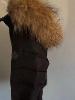 Svart pufferjacka med fuskpäls Hollies - Svart pufferjacka från Hollies med avtagbar huva och stor äkta päls i brunt. Jackan har dragkedja framtill och insida med leopardmönster. Perfekt för kalla vinterdagar/höst och riktigt snygg detalj med pälsen på huvan.