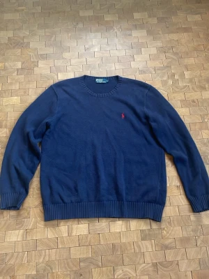 Mörkblå tröja från Polo Ralph Lauren - Klassisk mörkblå tröja från Polo Ralph Lauren med rund halsringning och ribbade muddar. Diskret röd logobrodyr på bröstet. Perfekt för en stilren look. Tröjan är sparsamt använd och den har inga defekter. 