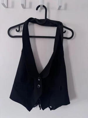 Svart väst med snörning och knappar - Snygg svart väst med djup v-ringning och bred krage. Västen har två knappar framtill och snörning bak som ger lite korsett vibes.