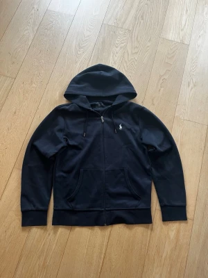 Ralph Lauren Zip Hoodie - Klassisk svart hoodie med dragkedja från Polo Ralph Lauren. Hoodien har huva med dragsko, två fickor framtill och den ikoniska vita Polo-loggan broderad på bröstet. Tillverkad i mjukt bomull. 100% äkta från ralph lauren, nypris ca 2000. Storlek S-M