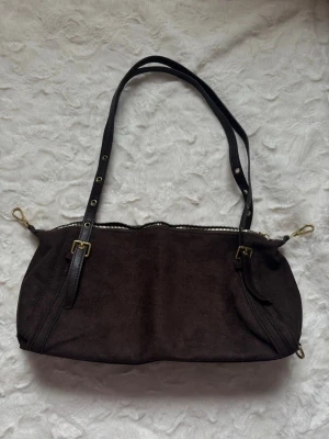 Mörkbrun axelväska i mocka - Snygg mörkbrun handväska i mocka med justerbara svarta axelremmar i läder och guldfärgade metalldetaljer. Väskan har dragkedja upptill och en avlång, mjuk form. Perfekt för dig som gillar stilrena accessoarer med en touch av vintage.
