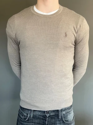 Beige stickad tröja från Ralph Lauren - Säljer en stilren beige stickad tröja från Ralph Lauren med rund halsringning och diskret broderad logga på bröstet. Tröjan är tunnstickad och har långa ärmar samt ribbade muddar vid ärmslut och nederkant. Perfekt till jeans eller chinos. Helt ny. Nypris 2299kr