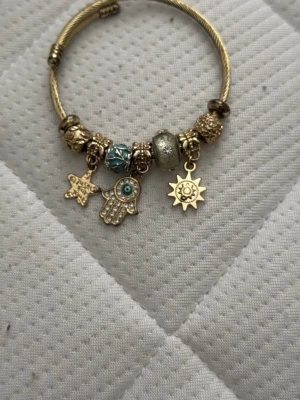 Guldigt armband med berlocker - Snyggt armband i guldfärgad metall med flera detaljerade berlocker, bland annat en stjärna, en hand med blå sten och en sol. Armbandet har pärlor i guld och turkos samt glittrande inslag. Perfekt accessoar för dig som gillar unika smycken.