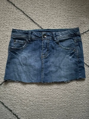 Blå jeanskjol med rå kant - Säljer en blå jeanskjol i klassisk denim med rå, avklippt kant. Kjolen har fem fickor, bälteshällor och knappstängning framtill. Perfekt för dig som gillar en avslappnad, vintage  och trendig stil.