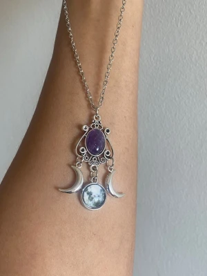 Silvrigt halsband med månberlocker - Unikt halsband i silverfärgad metall med en lila oval sten och detaljerade månberlocker. Hängsmycket har en bohemisk vibe med en rund bild av månen och två halvmånar. Perfekt för dig som gillar mystiska och magiska accessoarer.