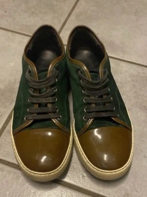 Gröna sneakers från Lanvin - Snygga sneakers från Lanvin i mörkgrönt tyg med bruna detaljer i läder och matchande bruna skosnören. Skorna har en vit sula och klassisk låg modell. Perfekta för dig som gillar stilrena och unika sneakers med exklusiv känsla.