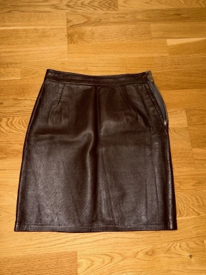 Svart skinnkjol med dragkedja - Snygg svart skinnkjol i kort modell med dragkedja i sidan och diskreta fickor fram. Kjolen har en stilren och klassisk design som passar perfekt till många olika looks. Materialet ger en cool vibe och den raka passformen gör den enkel att matcha.