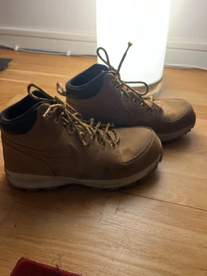 Bruna stövletter från Nike - Säljer ett par bruna stövletter från Nike med klassisk snörning och robust vit sula. Skorna har en högre modell med svart vadderad kant runt ankeln och är tillverkade i slitstarkt läder. Perfekta för dig som gillar en sportig och stilren look.