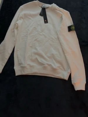 Vit sweatshirt från Stone Island - Säljer en stilren vit sweatshirt från Stone Island med den klassiska logotypen på vänster ärm. Tröjan har rund halsringning, ribbade muddar och är tillverkad i mjukt bomullsmaterial. Perfekt för en clean och avslappnad look.