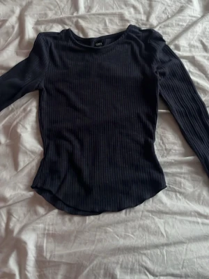 Svart ribbad långärmad topp från Only - En svart långärmad topp från Only med ribbad struktur och rund halsringning. Toppen har en slim passform och är perfekt att styla med jeans eller kjol. Enkel och stilren design som funkar till många olika looks.