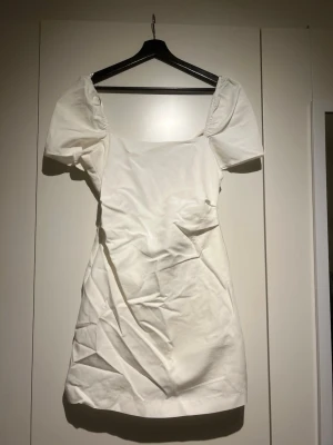 Vit kort klänning med puffärm - Säljer en vit kort klänning med puffärmar och fyrkantig urringning. Klänningen har en öppen rygg med dekorativ snörning och är tillverkad i ett bomullsliknande material. Perfekt för dig som gillar en feminin och trendig stil.