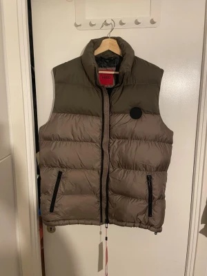 Dunväst från Hugo Boss i beige och oliv - Snygg dunväst från Hugo Boss i beige och olivgrönt med svart dragkedja och två fickor framtill. Västen har hög krage och innerfoder med Boss-logga. Perfekt för lager på lager och streetwear-vibe.