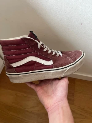 Vinröda Vans Sk8-Hi sneakers - Vinröda Vans Sk8-Hi sneakers med klassisk vit sidorand och vita snören. Ovandelen är i mocka och canvas, med vadderad ankel och vit gummisula. Perfekta för en streetwear-look och passar till många outfits.