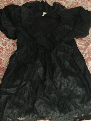 Svart volangklänning från Nelly - Supersnygg svart klänning från Nelly med volangdetaljer och v-ringad hals. Klänningen har korta puffärmar och är gjord i ett lätt, något glansigt material som ger en lyxig känsla. Perfekt för dig som vill sticka ut lite extra.