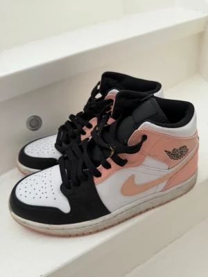 Air Jordan 1 Mid rosa och svart - Snygga Air Jordan 1 Mid sneakers från Nike i en fräsch färgkombination av rosa, svart och vitt. Skorna har klassisk Jordan-logga på sidan, perforerad tå och svarta skosnören. Materialet är en mix av läder och syntet. Perfekt för dig som vill sticka ut med din stil.