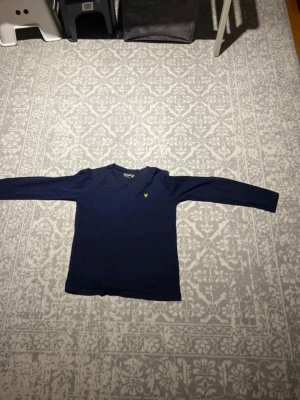 Marinblå långärmad topp Lyle & Scott - Snygg marinblå långärmad topp från Lyle & Scott med klassisk gul logga broderad på bröstet. Tillverkad i mjuk bomull som känns skön mot huden. Perfekt till jeans eller kjol för en clean och enkel stil.