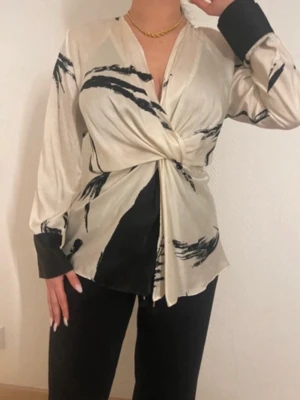 Beige mönstrad omlottblus med knut - Trendig beige omlottblus med svart abstrakt mönster och knut framtill. Långa ärmar med svarta muddar och guldknapp. Sidenliknande material som ger en lyxig känsla. Perfekt för att lyfta din outfit.
