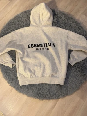 Grå Essentials Fear of God hoodie - Säljer en ljusgrå hoodie från Essentials Fear of God med svart logga på bröstet och stor text på ryggen. Hoodien har huva och magficka, samt ribbade muddar vid ärmar och nederkant. Perfekt för en avslappnad och trendig streetwear-look.