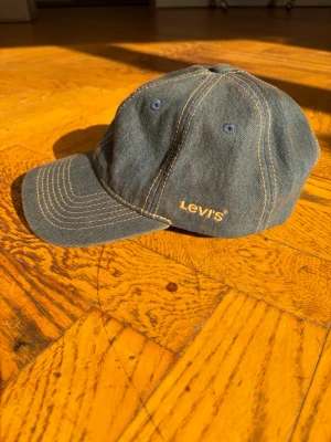 Blå denimkeps från Levi's - Säljer en klassisk blå denimkeps från Levi's med gul broderad logga på sidan. Kepsen har böjd skärm, synliga kontrastsömmar och metallhål för ventilation. Perfekt accessoar för en avslappnad streetwear-look.