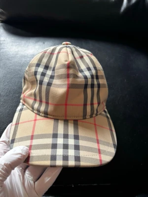 Beige keps med klassiskt rutmönster - Snygg beige keps från Burberry med det ikoniska rutmönstret i svart, vitt och rött. Kepsen har böjd skärm och justerbar passform. Perfekt accessoar för en trendig och stilren look.