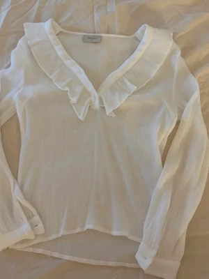 Vit volangblus  - Superfin vit blus med volangdetalj längs v-ringningen. Blusen har långärmade ärmar med knappar vid ärmsluten och är gjord i ett lätt, transparent tyg som ger en luftig känsla. Perfekt att styla med jeans eller kjol.