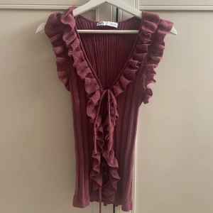 Lila volangtopp - Supersöt lila topp från Zara med djup v-ringning och volangdetaljer längs kanterna. Toppen är ärmlös och har knyt framtill för extra detalj. Perfekt för dig som vill sticka ut med en romantisk vibe.