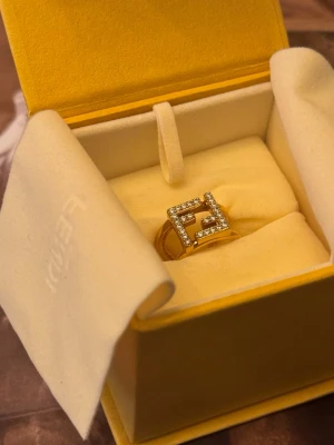 Fendi ring med kristaller - Säljer en exklusiv guldfärgad ring från Fendi med fyrkantig design och gnistrande kristaller runt hela Fendi-loggan. Ringen levereras i original ask och har en modern, lyxig känsla som verkligen sticker ut.