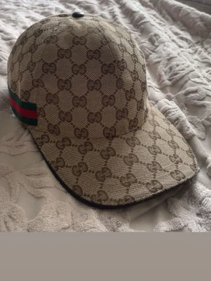 Beige keps från Gucci med GG-mönster - Säljer en beige keps från Gucci med det klassiska GG-monogrammet över hela kepsen. På sidan finns en grön och röd rand som signaturdetalj. Kepsen har justerbart band bak och är tillverkad i canvasmaterial med mörkbrunt innerfoder.