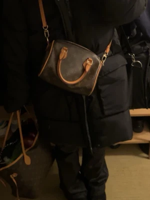 Brun väska  - Säljer en klassisk  handväska i brunt monogrammönster med beige och ljusbruna detaljer. Väskan har både axelrem och två små handtag, samt guldfärgade metalldetaljer. Perfekt storlek för det viktigaste och en ikonisk accessoar. Den har en liten fläck som inte jag kan få bort. Den är ganska stor man får plats med mycket saker.