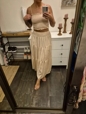 Beige set, smockad linnetopp + kjol - Beige set om kjol och topp i mjukt, luftigt material. Kjolen har smockad midja och är stretchig och följsam, toppen är likadan med justerbara axelband. Storlek S/M men kan även passa XS, kjolens total längd är ca 90 cm (jag är 164) Köpt på Cypern. Endast använd vid ett tillfälle så skicket är som nytt utan skavanker.