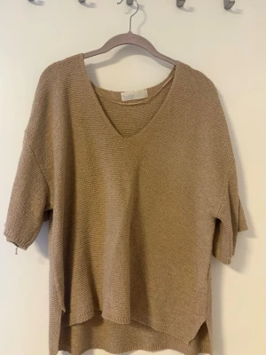 Beige stickad t-shirt från Samsoe Samsoe - Beige stickad t-shirt från Samsoe Samsoe med v-ringning och korta ärmar. Tröjan har en avslappnad passform och är tillverkad i ett mjukt, tunt stickat material. 