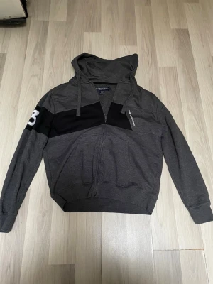 Mörkgrå hoodie från polo - Snygg mörkgrå hoodie med dragkedja från U.S. Polo Assn. Har svart panel över bröstet och en vit siffra på ärmen. Tillverkad i bomull och polyester, med huva och dragsko. Perfekt för en avslappnad och sportig stil.