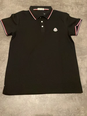 Svart polotopp från Moncler - Svart polotopp från Moncler med klassisk krage och knappar framtill. Toppens krage och ärmslut har snygga röda och vita detaljer. Moncler-logga på bröstet. Perfekt för dig som gillar stilrena plagg med exklusiv känsla.